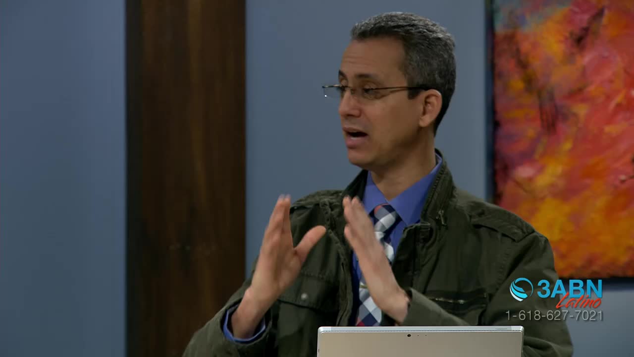 A man in glasses gestures with both hands, his fingers spread wide as he speaks. He's wearing a jacket and a patterned tie, and a laptop sits in front of him.
