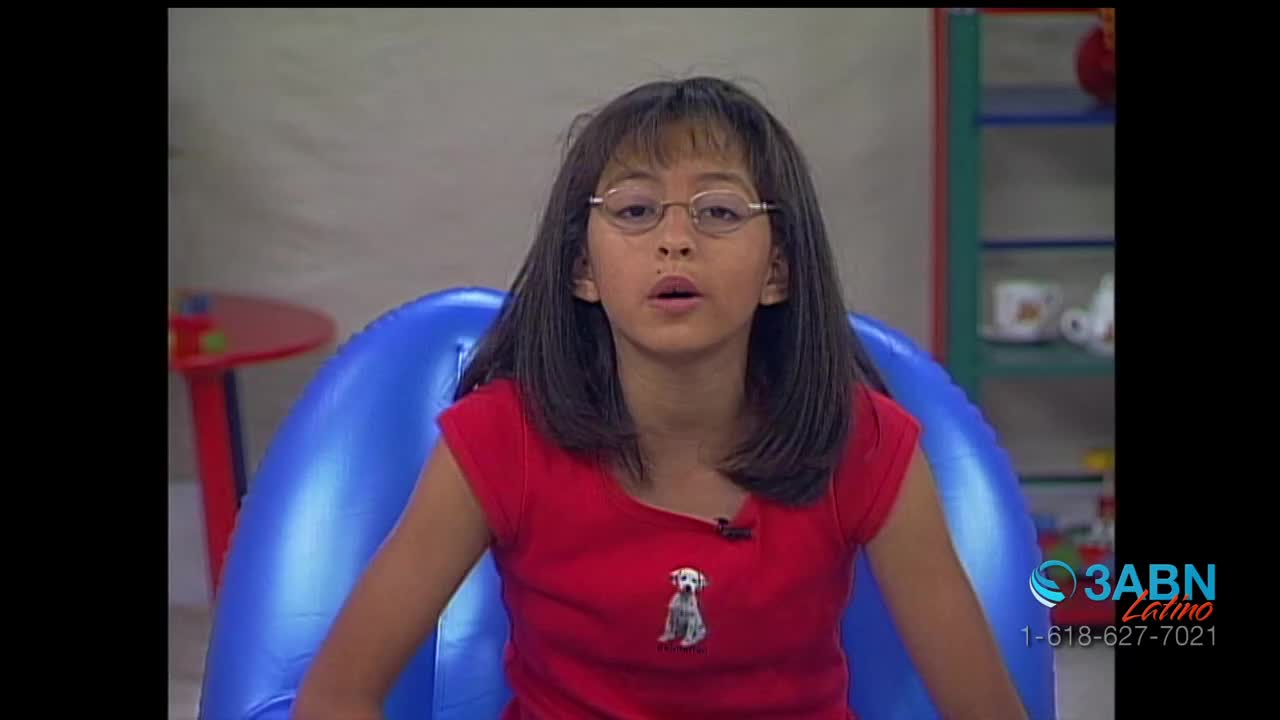 A young girl in a red shirt, wearing glasses, is speaking into a microphone. She's seated in a blue inflatable chair, likely on a set for 3ABN Latino.

