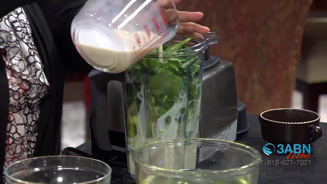 A hand is pouring a stream of light-colored liquid from a measuring cup into a blender filled with spinach. The scene is part of a 3ABN Latino broadcast from the United States.
