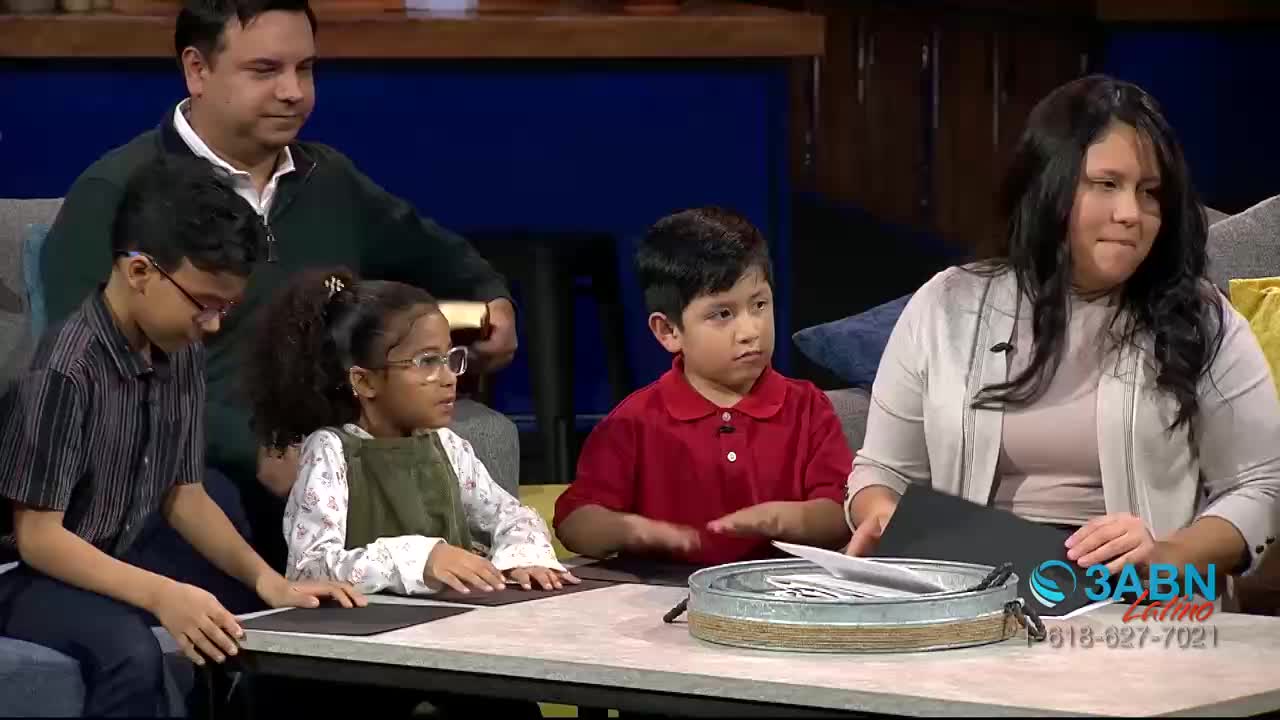 A family sits around a table, the children looking toward the camera. The woman on the right holds a black card, and the 3ABN Latino logo is visible.
