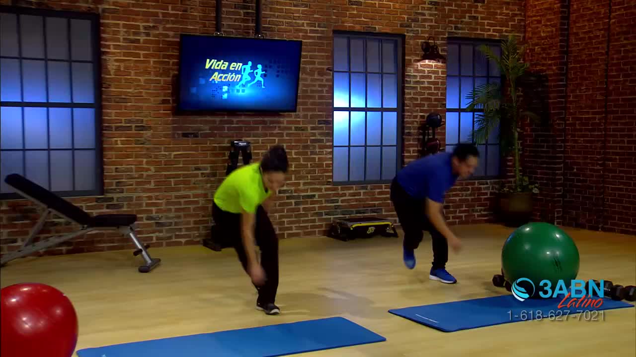 Two people are doing exercises on mats, moving in unison. The 3ABN Latino studio has a brick wall backdrop and a screen displaying "Vida en Acción."
