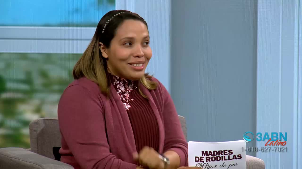 A woman with a headband smiles, her maroon cardigan open over a patterned shirt. A paper with Spanish text and the 3ABN Latino logo rests on the table before her.