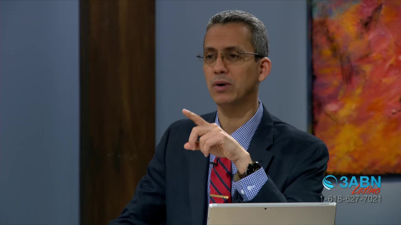 A man in a suit gestures with his right hand while speaking. He's on a set that includes a colorful abstract painting and a logo for 3ABN Latino.