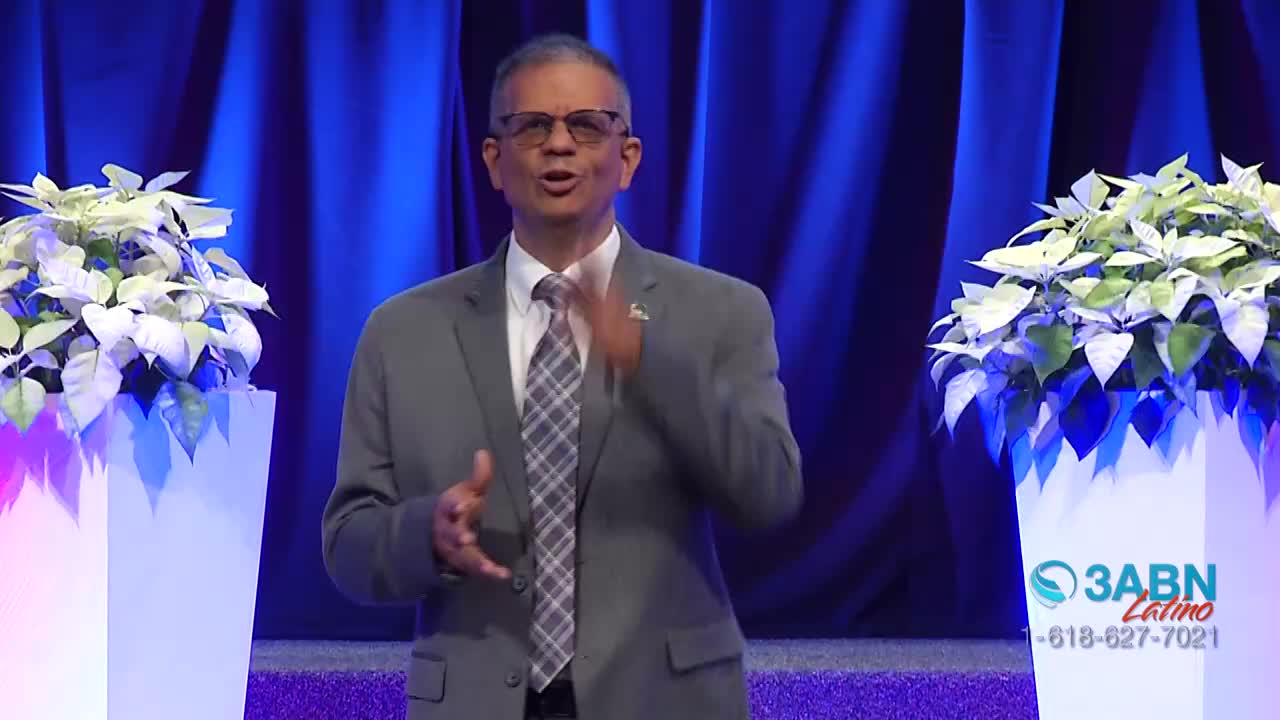 A man in a gray suit speaks with animated hand gestures. White poinsettias in tall planters flank him, their leaves catching the light.