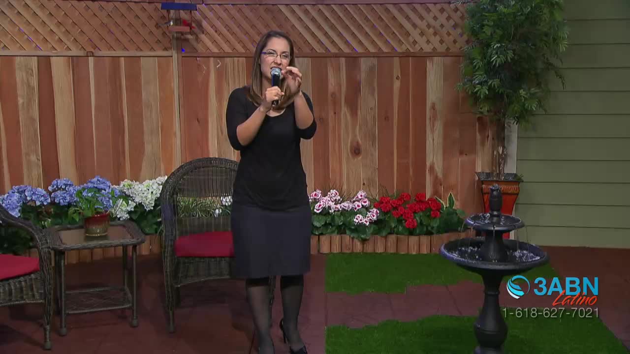 A woman holds a microphone and gestures with her hands while speaking. Colorful flowers bloom behind her, and a small fountain trickles water.