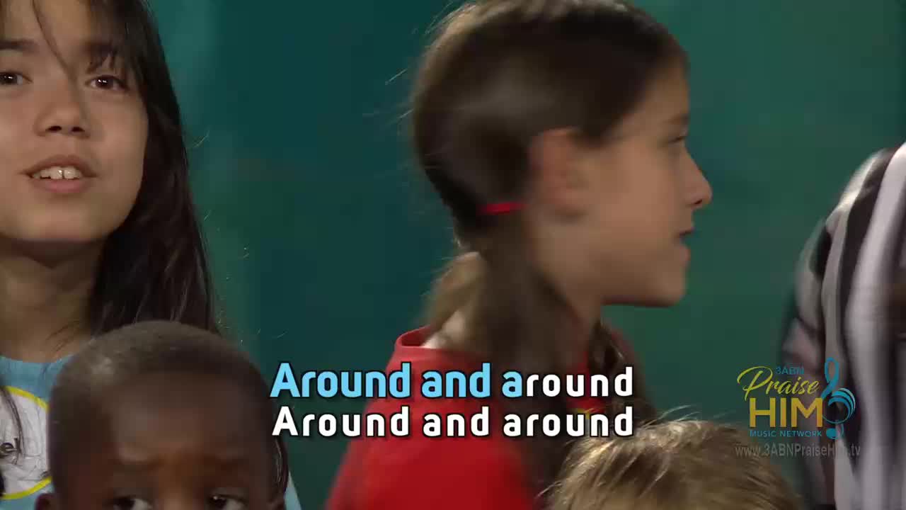 Children are singing, their faces lit up as they look to the right. The words "Around and around" are displayed on screen, and the logo for 3ABN Praise Him Music Network is visible.
