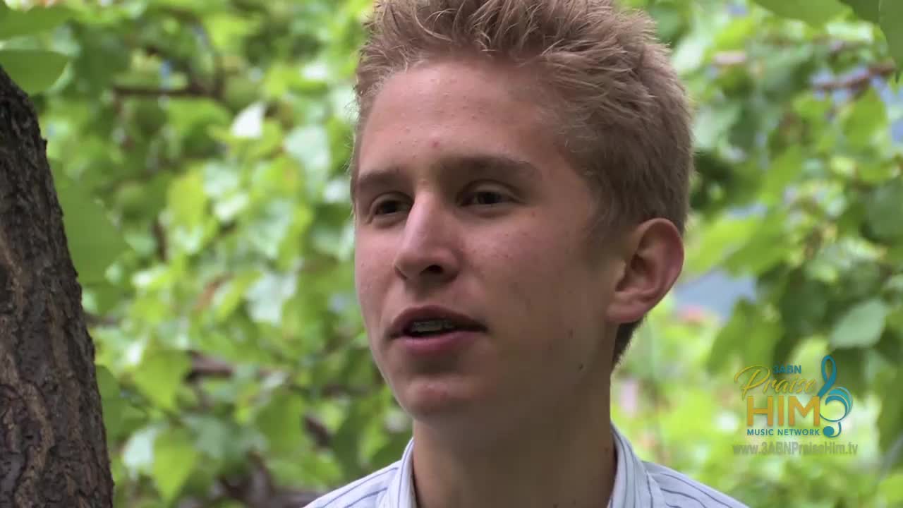 A young man with braces is speaking, his gaze directed off-camera. Behind him, green leaves and a tree trunk provide a natural backdrop for the 3ABN Praise Him Music Network logo.
