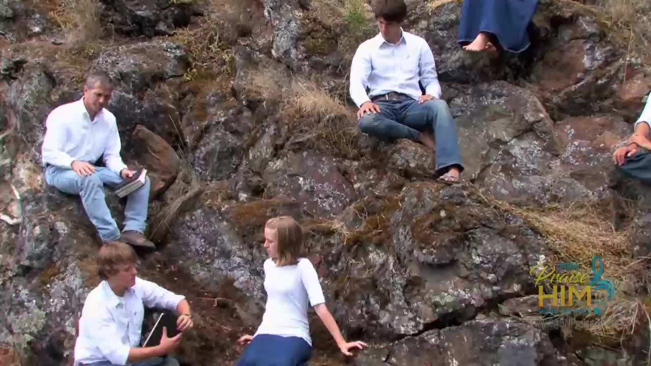 A group of people in white shirts and jeans are sitting on a rocky hillside. Some of them hold books and appear to be part of a production for the 3ABN Praise Him Music Network.
