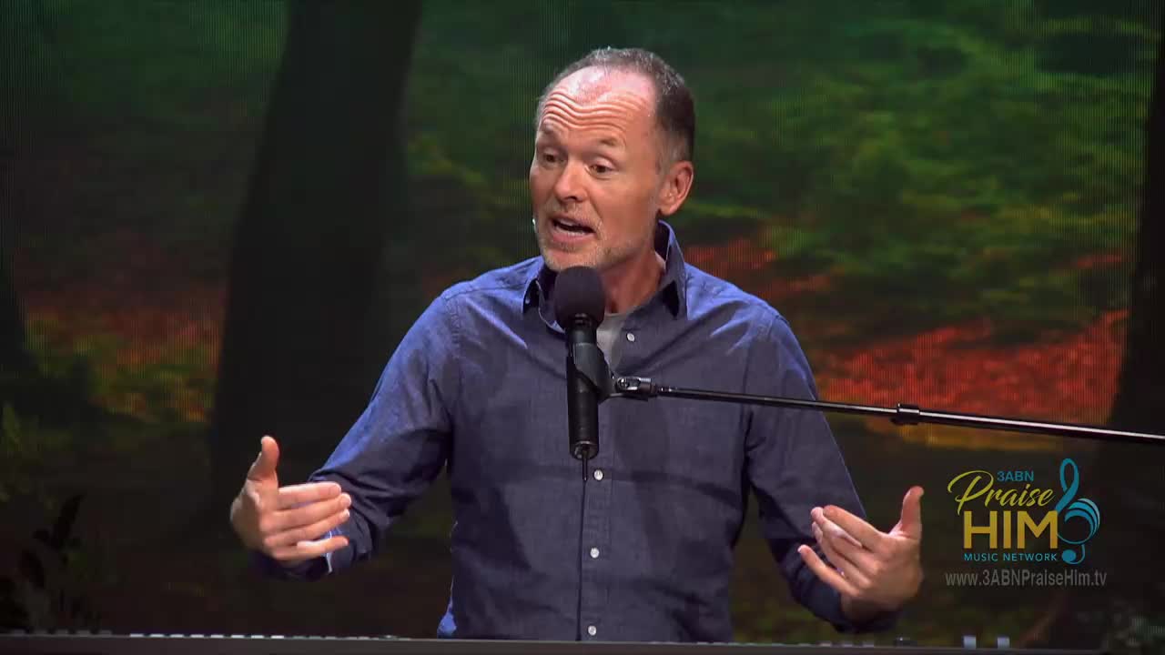 A man in a blue shirt gestures with both hands, speaking into a microphone. The 3ABN Praise Him Music Network logo is visible in the corner.
