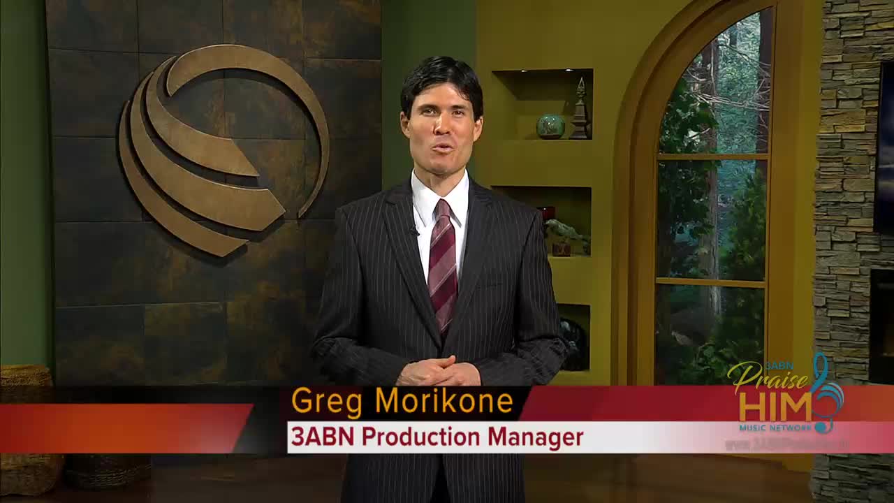 Greg Morikone, the 3ABN Production Manager, stands in a studio, speaking. A lower third identifies him as he talks, with the "Praise Him" logo visible in the corner.
