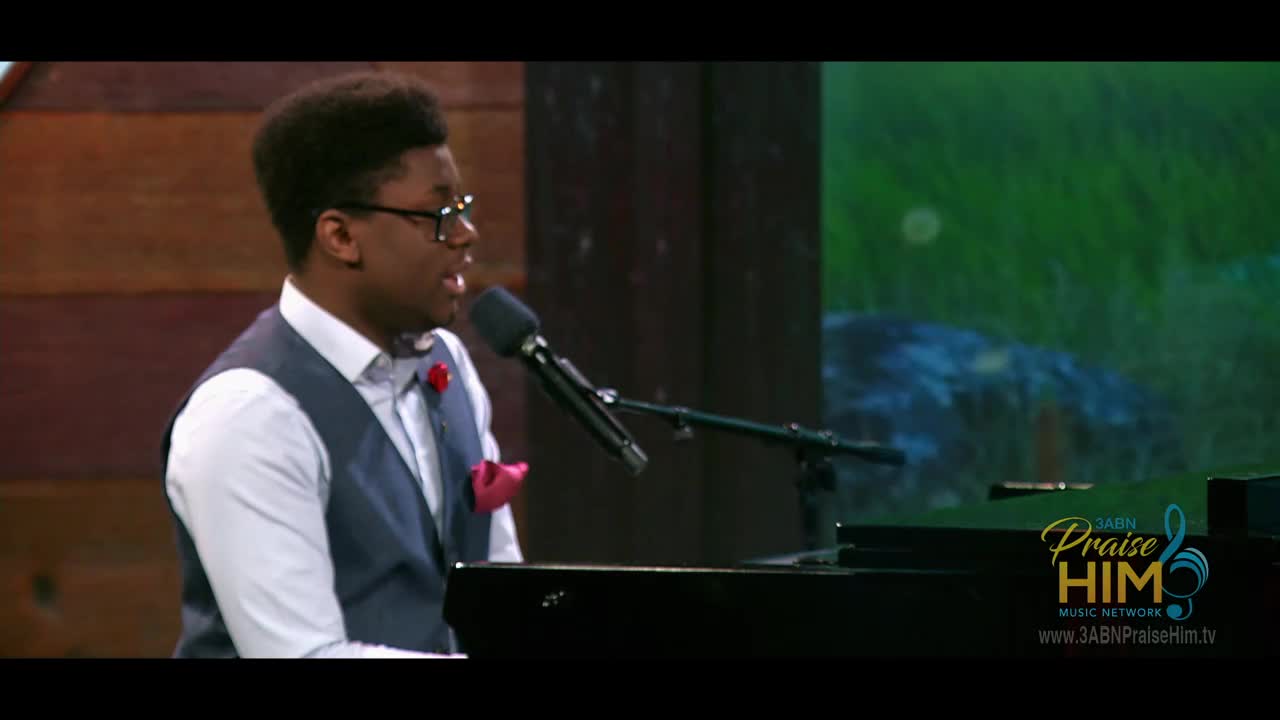 A young man in a vest and bow tie sings into a microphone while playing a piano. The 3ABN Praise Him Music Network logo is visible in the corner.
