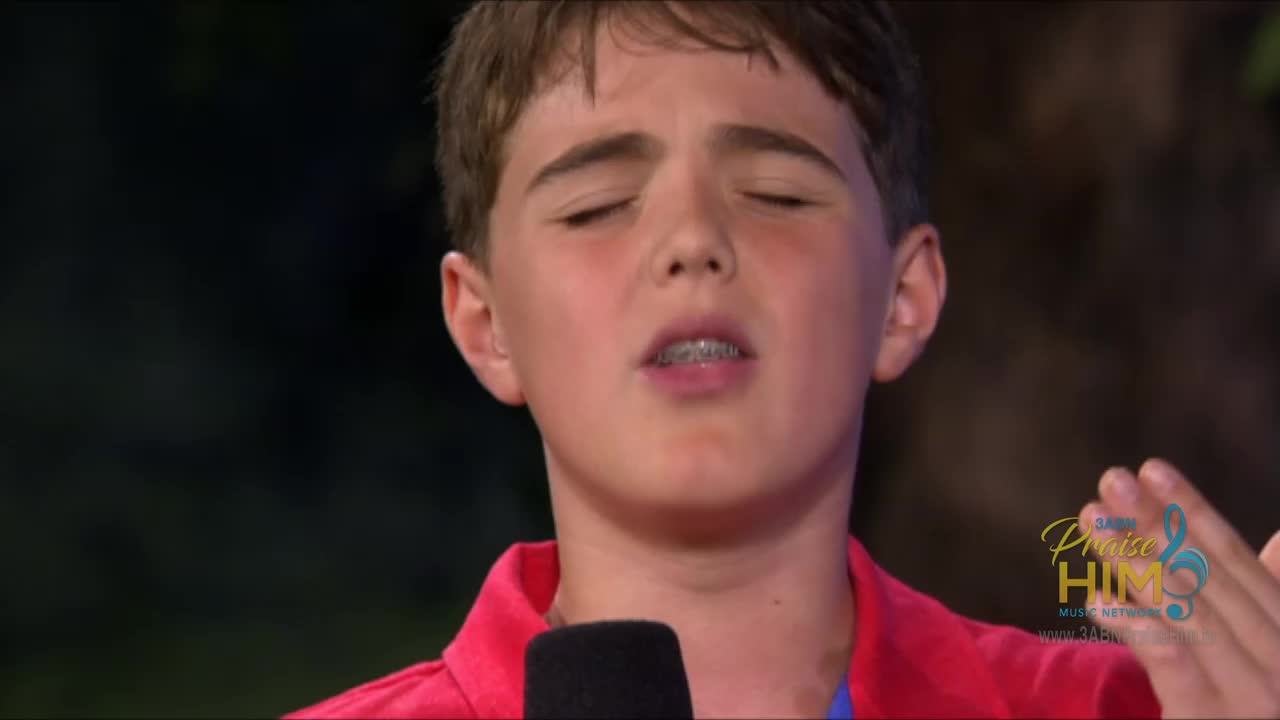 A young boy in a red shirt sings with his eyes closed, a microphone close to his mouth. The "Praise Him Music Network" logo is visible, indicating a performance on the 3ABN network in the United States.
