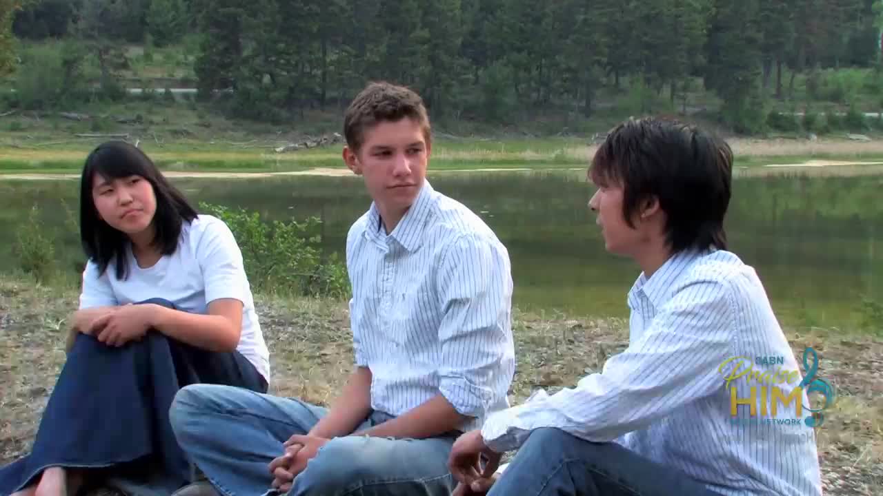 Three young people sit near a lake, engaged in a conversation. The girl in the white shirt and dark skirt looks toward the other two, while the boys in blue jeans and striped shirts seem to be talking to each other.
