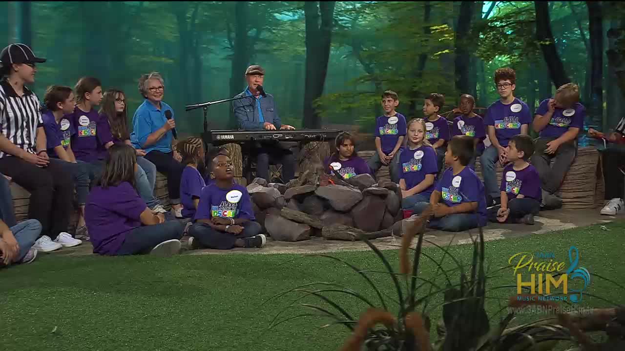 A man plays a keyboard as children in purple shirts sit around a pile of rocks. The scene is part of a broadcast from the 3ABN Praise Him Music Network, with a backdrop of trees.
