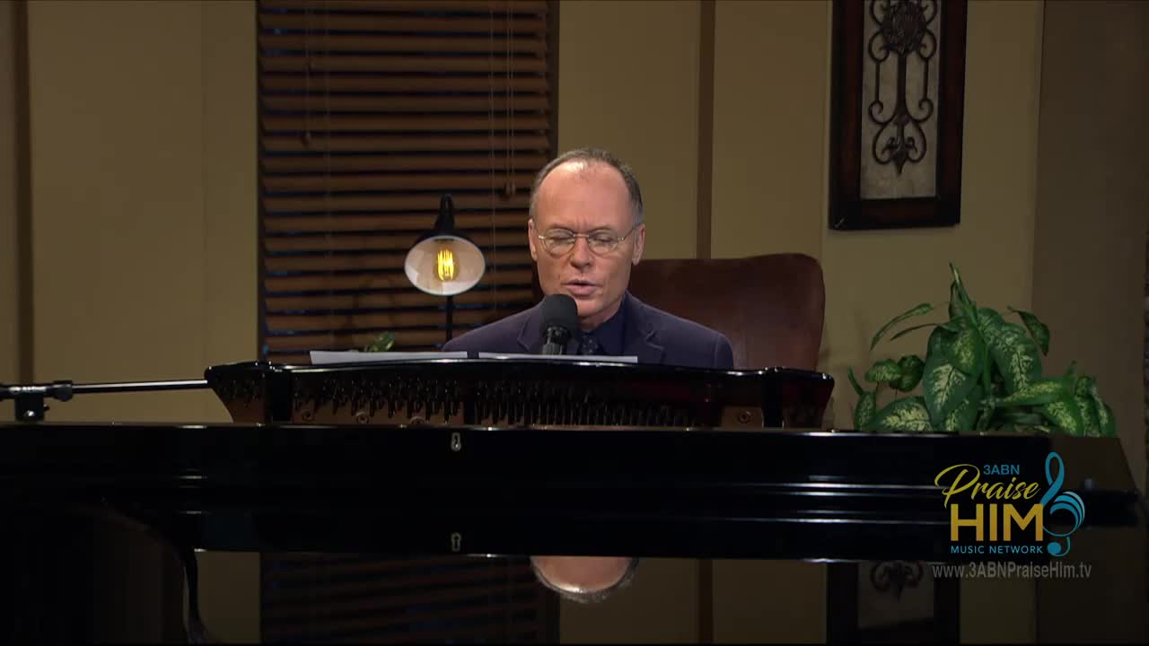A man in a dark suit sits at a grand piano, speaking into a microphone. The 3ABN Praise Him Music Network logo is visible in the corner.
