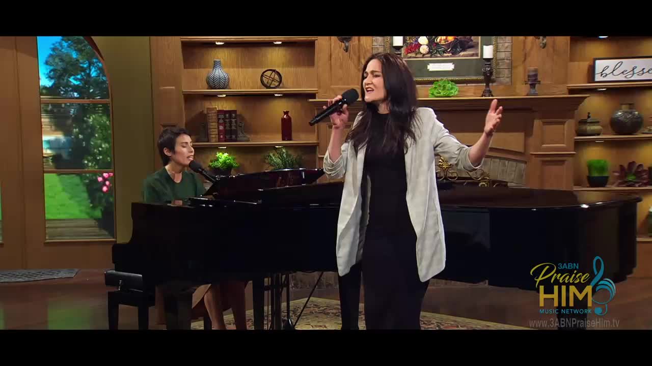 A woman in a black dress and light jacket sings into a microphone, her arm raised. Beside her, another woman plays a grand piano on a set for 3ABN Praise Him Music Network.
