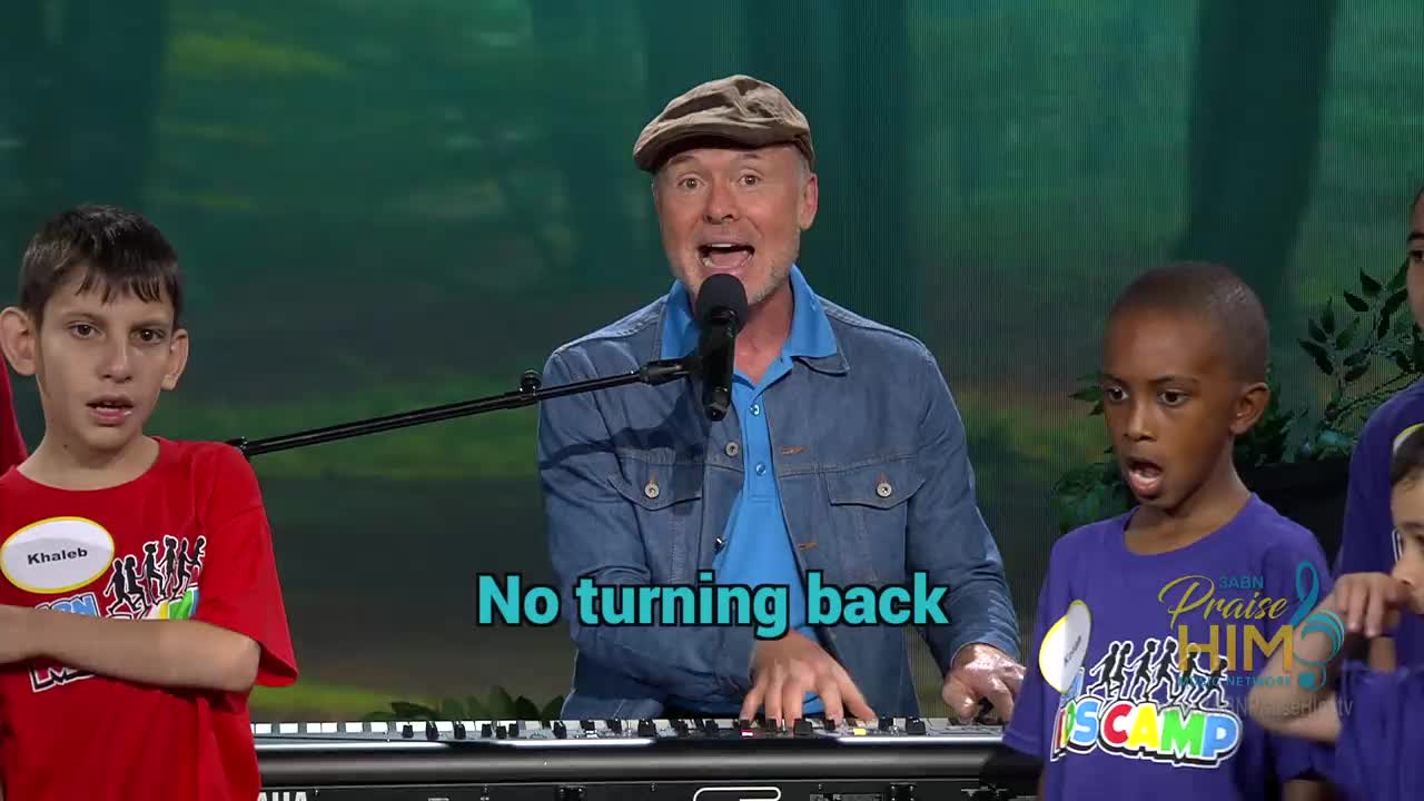 A man in a denim jacket sings into a microphone, playing a keyboard with a group of children beside him. The children, some wearing "Praise Him" shirts, appear to be singing along on the 3ABN Praise Him Music Network.
