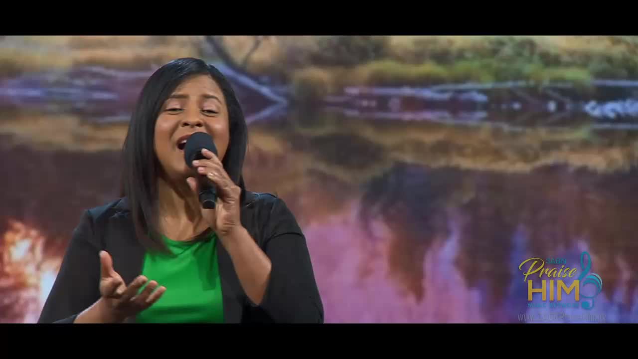 A woman, holding a microphone, sings with her hands open, and her eyes closed. Behind her, a blurred backdrop of a landscape is visible, with the words "Praise Him" in the bottom right corner.
