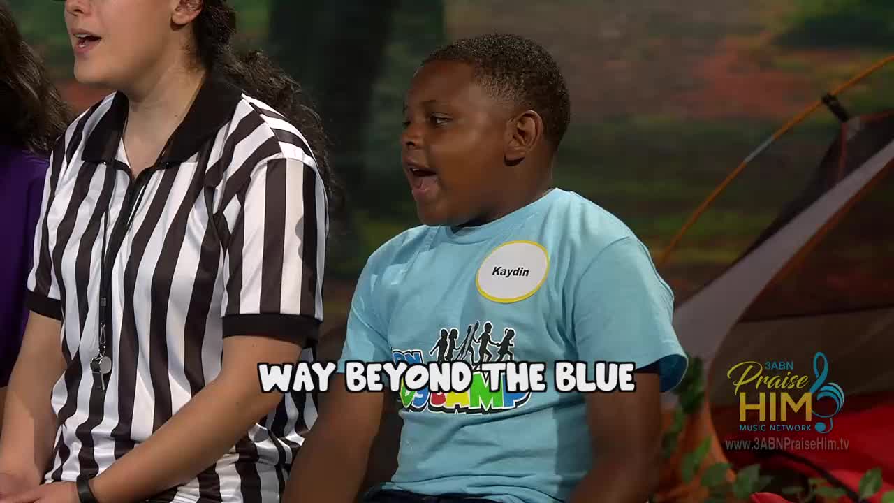 A young boy wearing a light blue shirt with "Camp" printed on it is singing, mouth open. Beside him, a person in a black and white striped referee shirt appears to be singing as well, on a set for 3ABN Praise Him Music Network.
