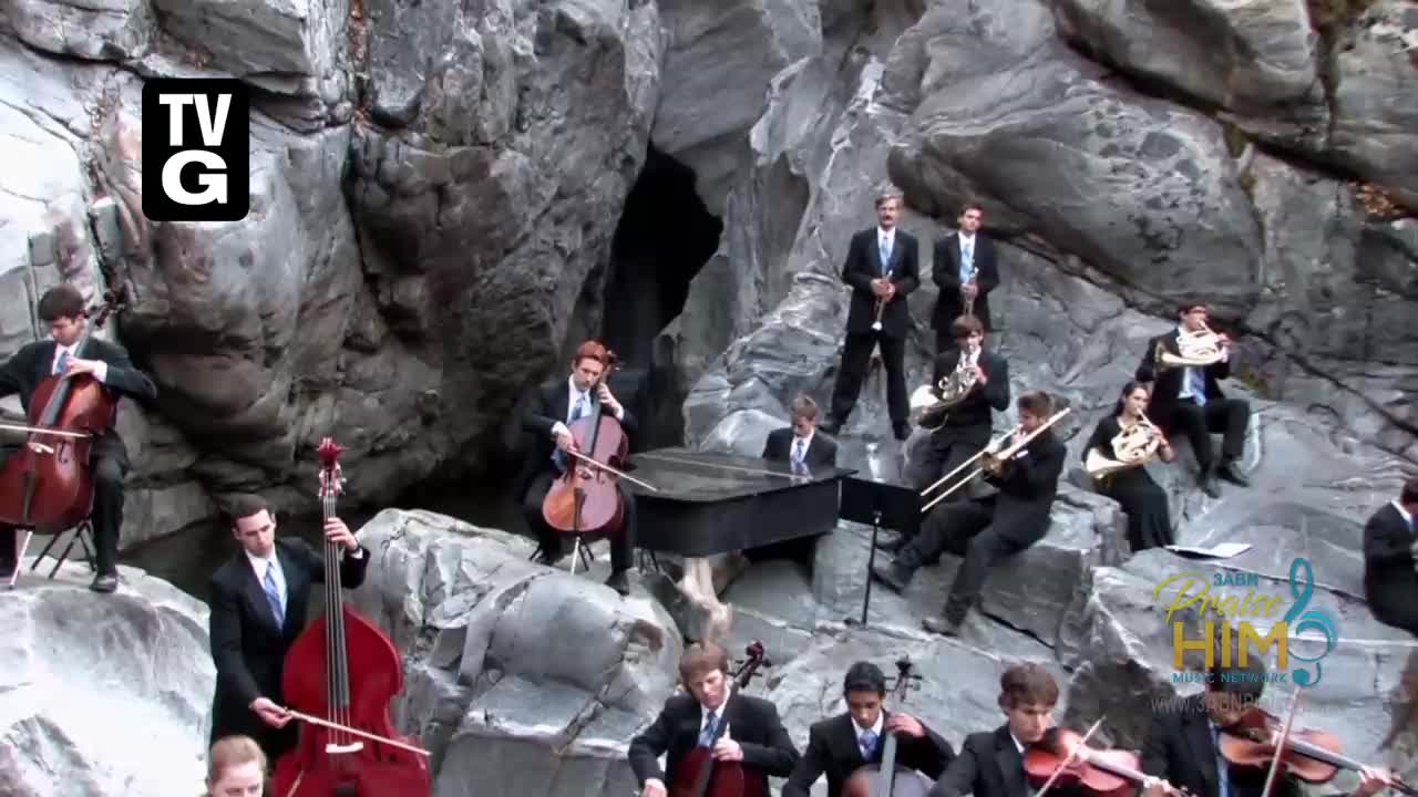 An orchestra, dressed in suits, is arranged on a rocky hillside. The group, likely from the 3ABN Praise Him Music Network, is playing in front of a cave entrance.

