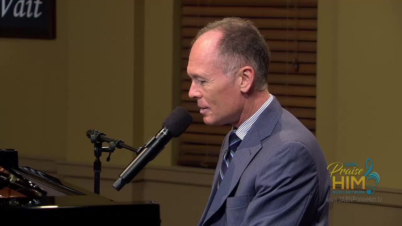 A man in a blue suit sits at a piano, speaking into a microphone. The 3ABN Praise Him Music Network logo is visible on the right side of the screen.
