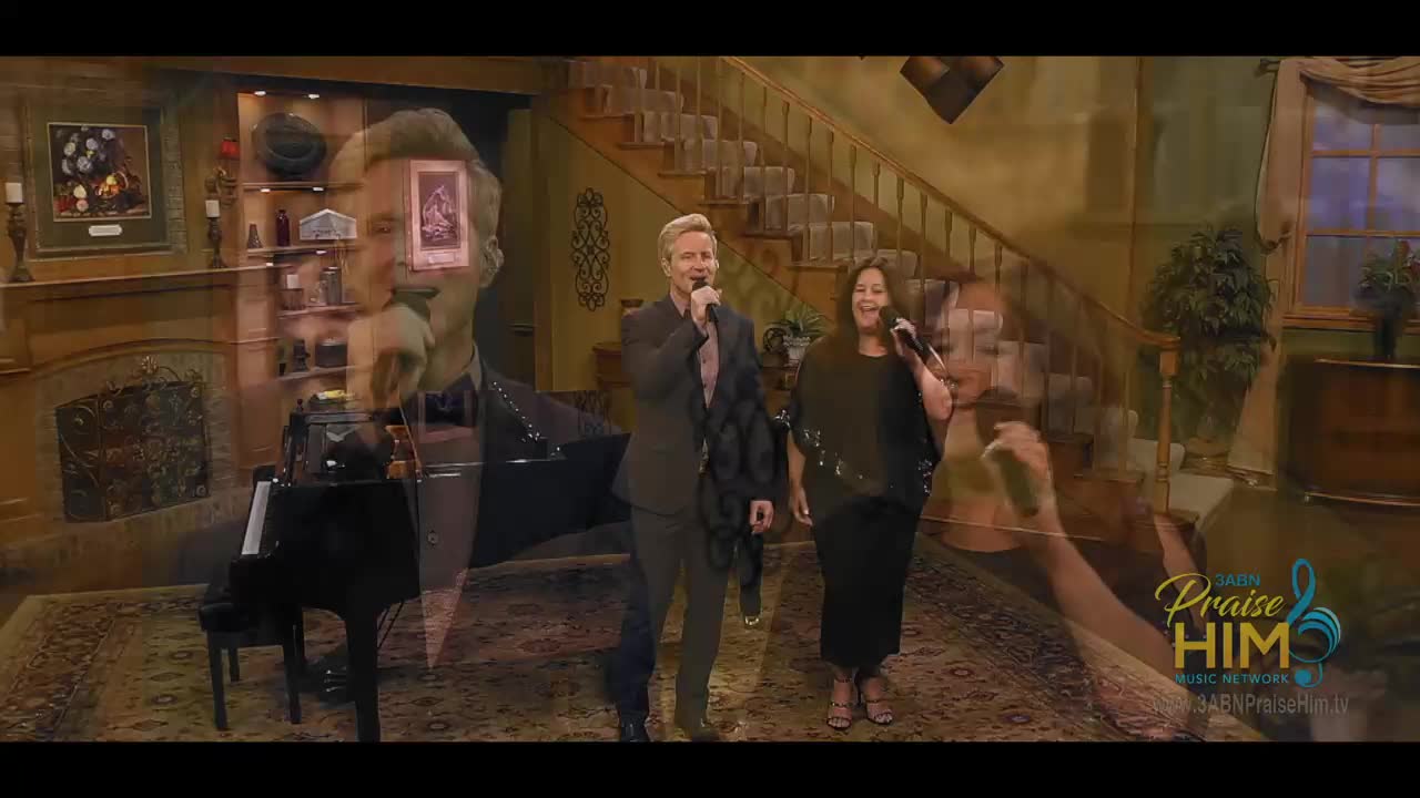 A man and a woman, microphones in hand, sing on a set for 3ABN Praise Him Music Network. The background includes a piano, a staircase, and a fireplace, all suggesting a home in the United States.
