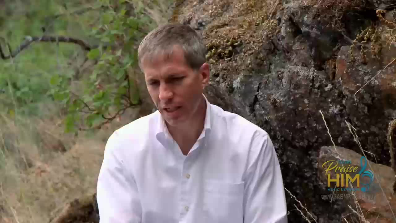A man in a white shirt speaks, likely for 3ABN Praise Him Music Network, in the United States. He sits outside near a rocky hillside.
