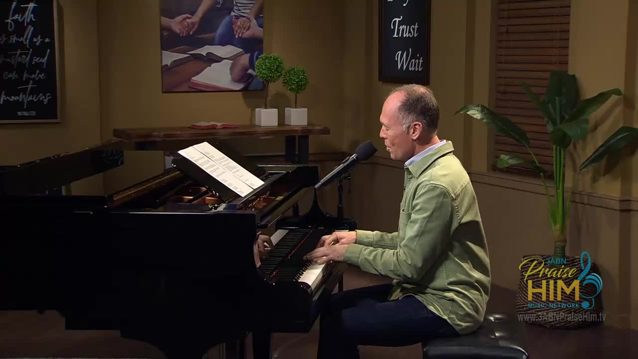 A man in a green shirt sits at a grand piano, his fingers moving over the keys. He's singing into a microphone, part of a broadcast on 3ABN Praise Him Music Network.
