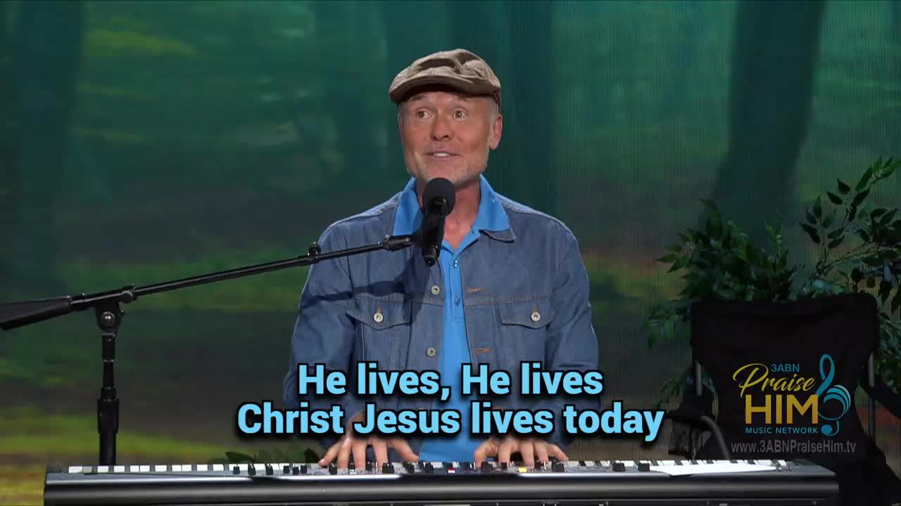 A man in a denim jacket and cap sings into a microphone, his fingers dancing across the keys of a keyboard. The words "He lives, He lives, Christ Jesus lives today" are displayed on the screen, likely part of a broadcast from 3ABN Praise Him Music Network.
