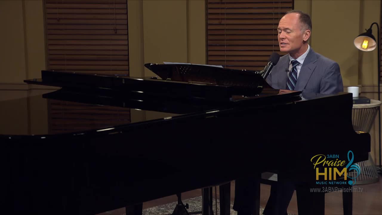 A man in a suit sings into a microphone while seated at a grand piano. The 3ABN Praise Him Music Network logo is visible on the lower right.
