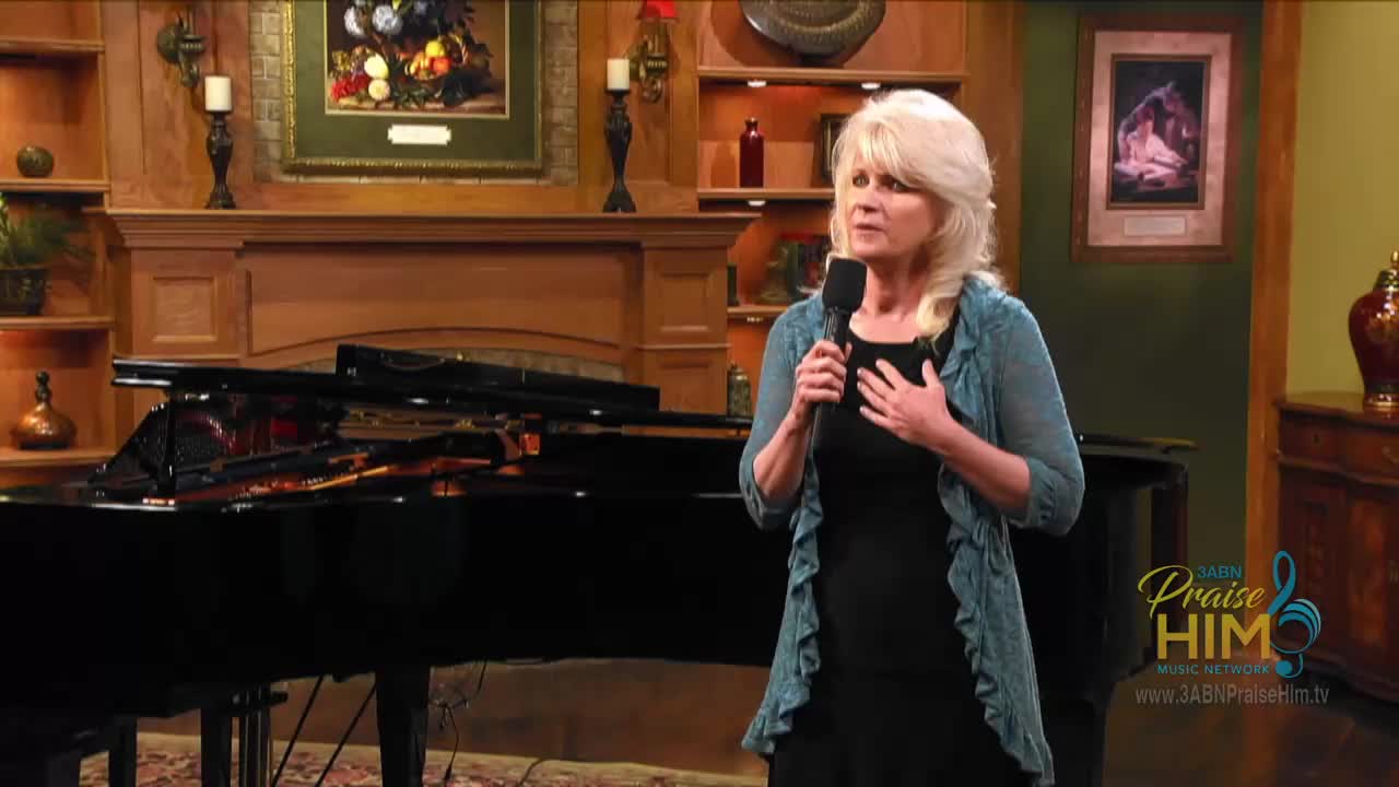 A woman stands on a set, holding a microphone and speaking, her hand resting on her chest. Behind her is a grand piano and a logo for the 3ABN Praise Him Music Network.
