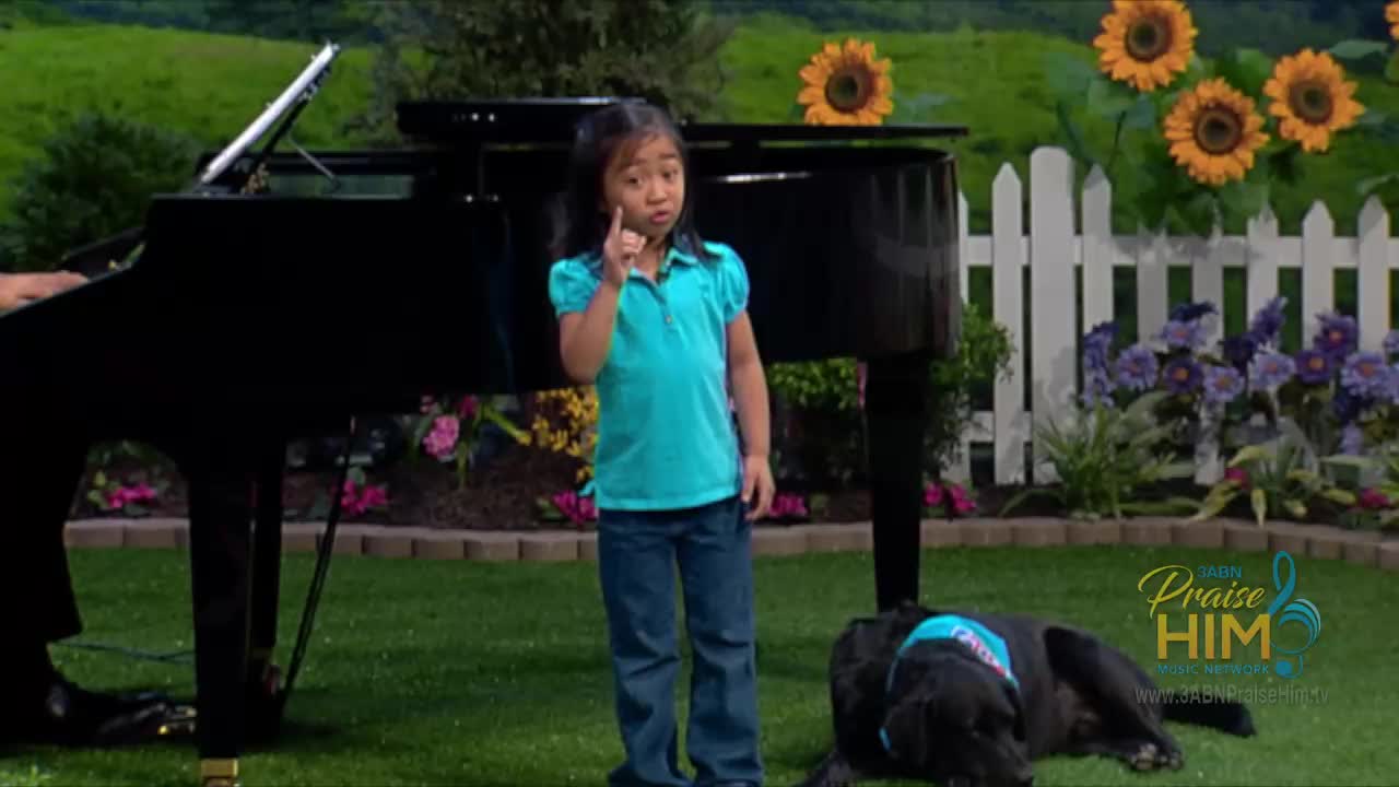 A young girl, wearing a turquoise shirt, stands in front of a grand piano, pointing her index finger upward. A black dog rests on the grass nearby, while sunflowers and a white picket fence decorate the background of this 3ABN Praise Him Music Network scene.
