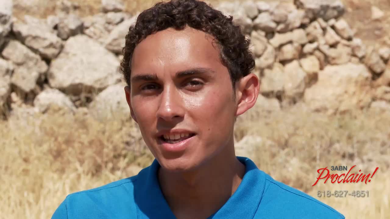 A young man in a blue shirt is speaking, his face lit by the sun. Behind him, a stone wall and the 3ABN Proclaim! logo are visible.

