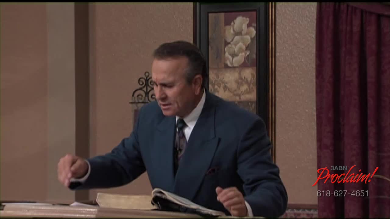 A man in a dark suit gestures emphatically while speaking. He is standing behind a lectern, with a Bible open in front of him, in a studio for the 3ABN Proclaim! Network in the United States.
