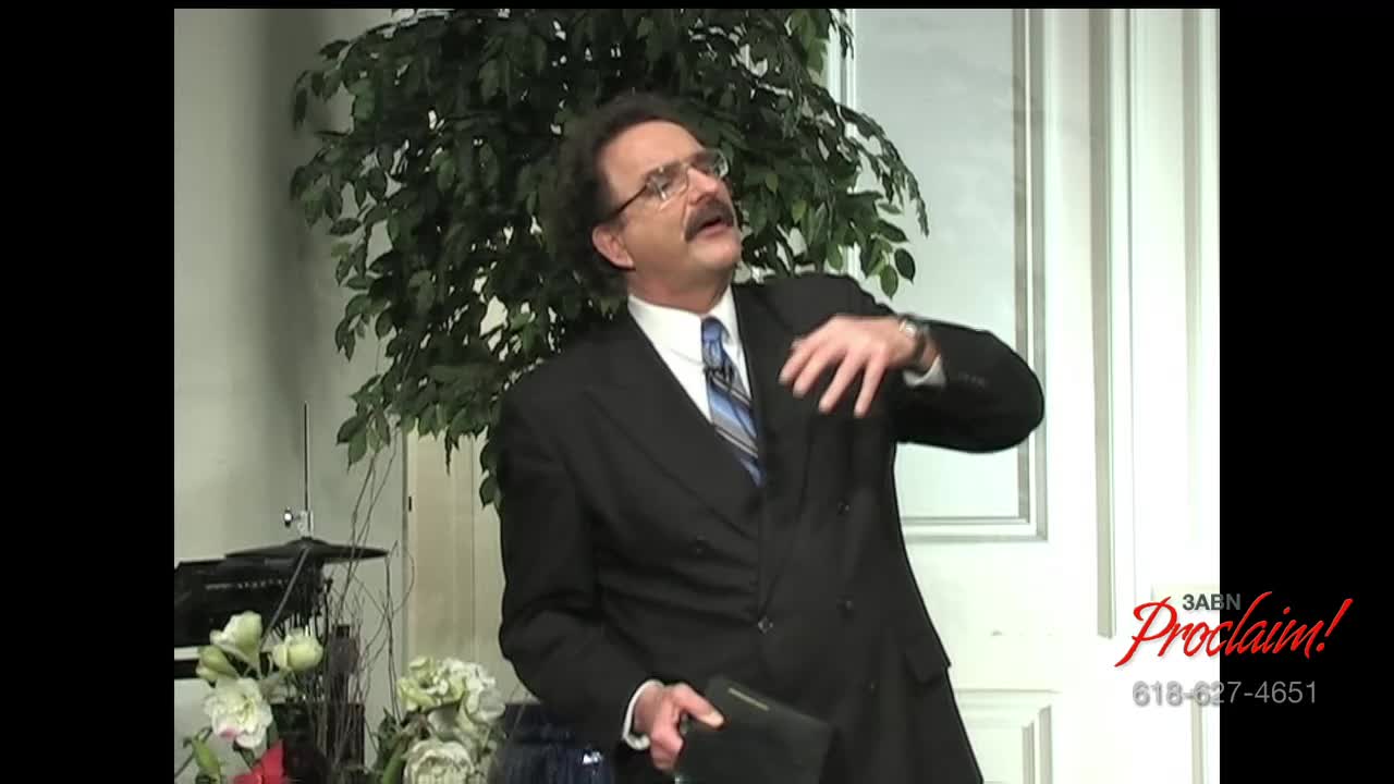 The man in the suit gestures with his right hand, his eyes lifted towards the ceiling as he speaks.  A small, black book rests in his left hand, and the 3ABN Proclaim! logo is visible.
