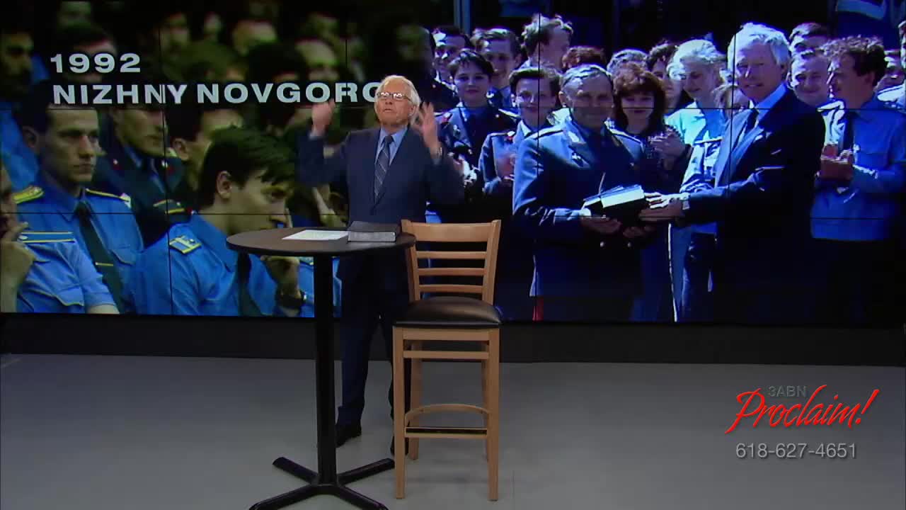 A man in a suit gestures with both hands in front of a large screen showing a presentation about Nizhny Novgorod in 1992. Behind him, a table and chair are set up, and the 3ABN Proclaim! Network logo is visible.
