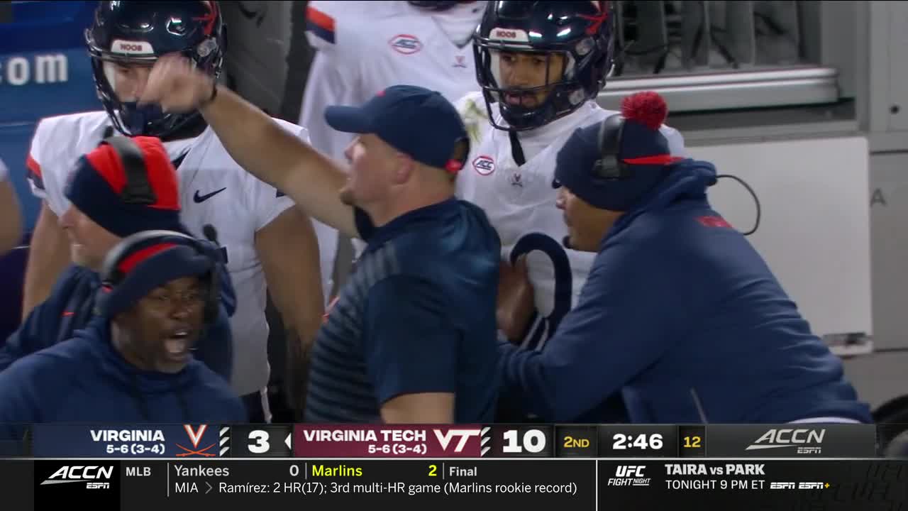 The Virginia players and coaches are huddled on the sideline, animated. A coach in a navy blue cap gestures emphatically toward the field.
