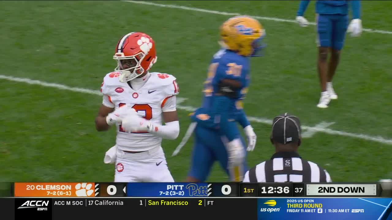 A Clemson player in orange and white is jogging forward, looking towards the sideline. A Pitt player in blue and gold is moving in the opposite direction, with the referee watching nearby.
