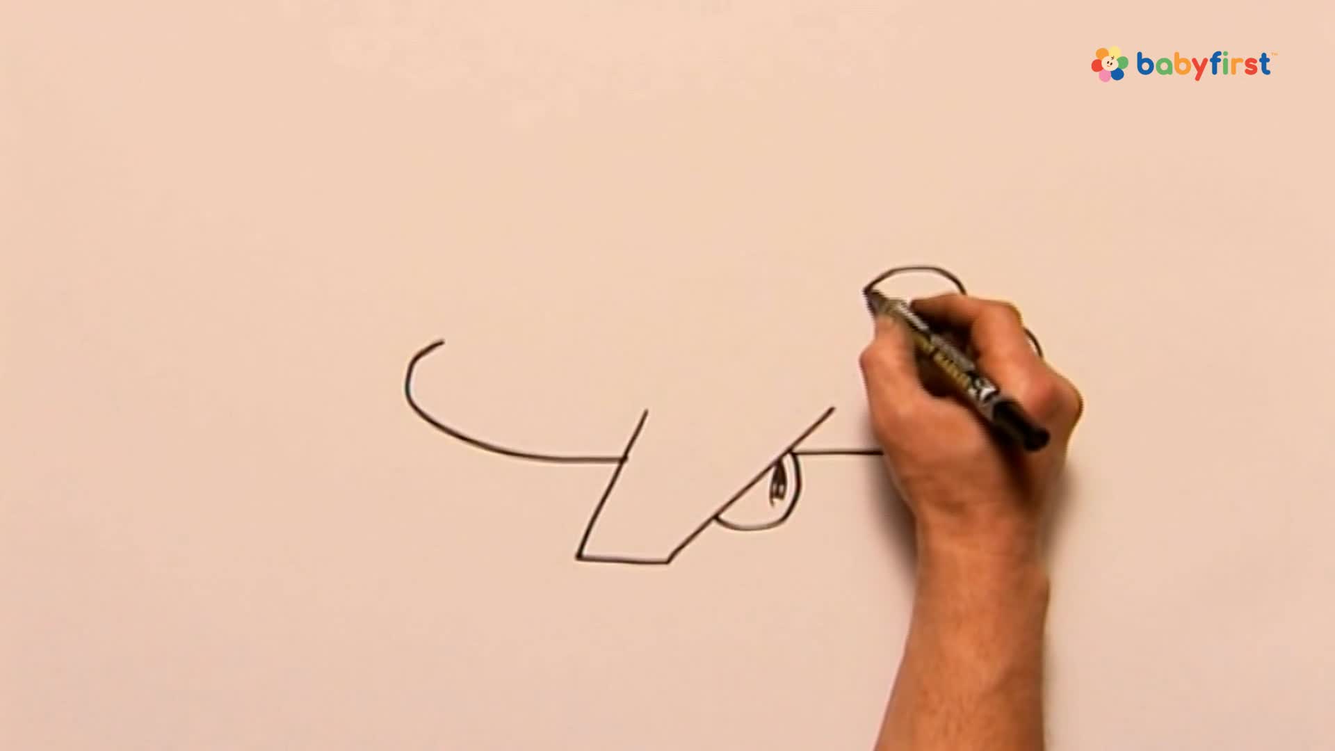 A hand holds a black marker, adding a curved line to the top of a drawing. The sketch of a cartoon face is taking shape on a white surface.
