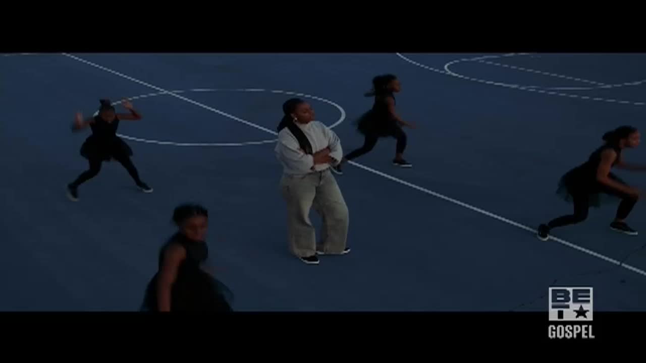 A woman in a sweatshirt and jeans stands with arms crossed, watching young girls in black dresses dance on a basketball court. They move with energy, their arms and legs in motion as they perform.
