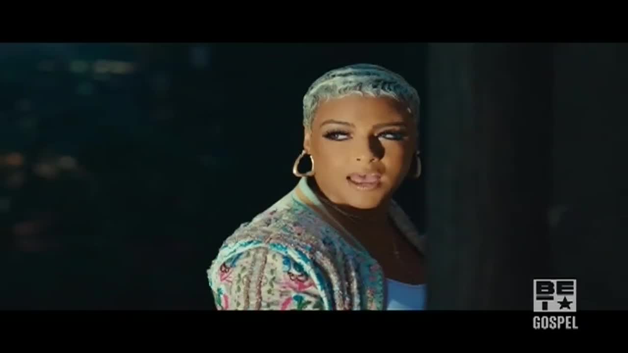 A woman with short, wavy hair peeks out from a dark space, her expression intense. She's wearing a colorful jacket and hoop earrings, and the BET Gospel logo is visible in the corner.
