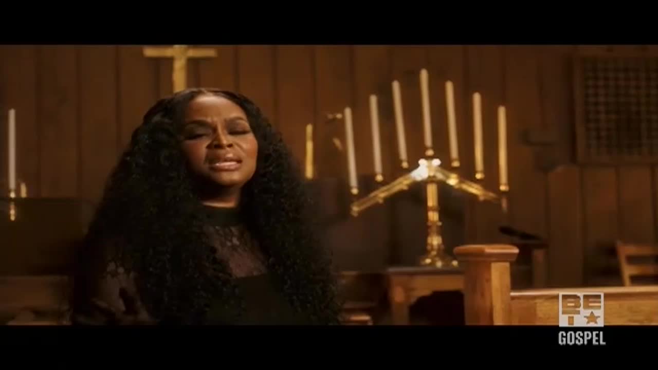 A woman with long, dark hair sings with her eyes closed, the emotion evident on her face. The set, a church interior, is lit warmly, with candles and a cross visible in the background.
