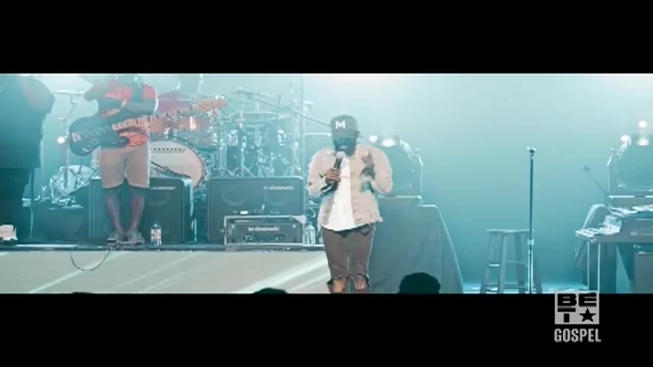 A man in a denim jacket stands center stage, speaking into a microphone. Behind him, a band is set up, ready to play on the BET Gospel stage.
