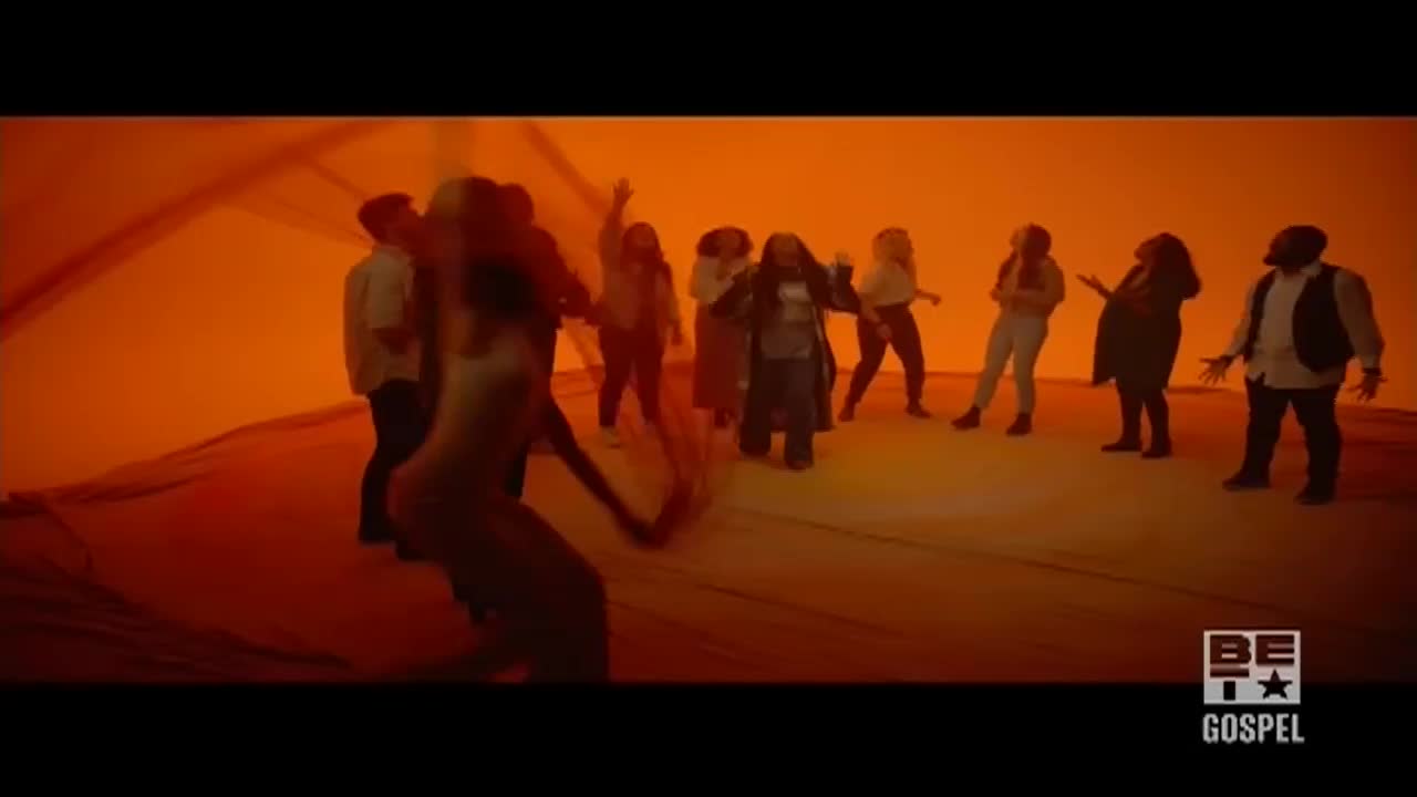 A group of people are singing and dancing in front of a bright orange backdrop, with a woman in the center wearing a long coat. The BET Gospel logo is visible in the bottom right corner.
