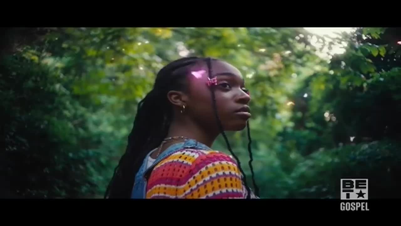 A young woman with long braids gazes off to the side, a pink butterfly clip in her hair. She's wearing a colorful sweater, and the BET Gospel logo sits in the corner.
