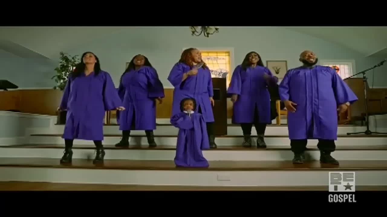A gospel choir, dressed in matching purple robes, stands on a stage. A small child, also in a robe, stands centered in front of the group.
