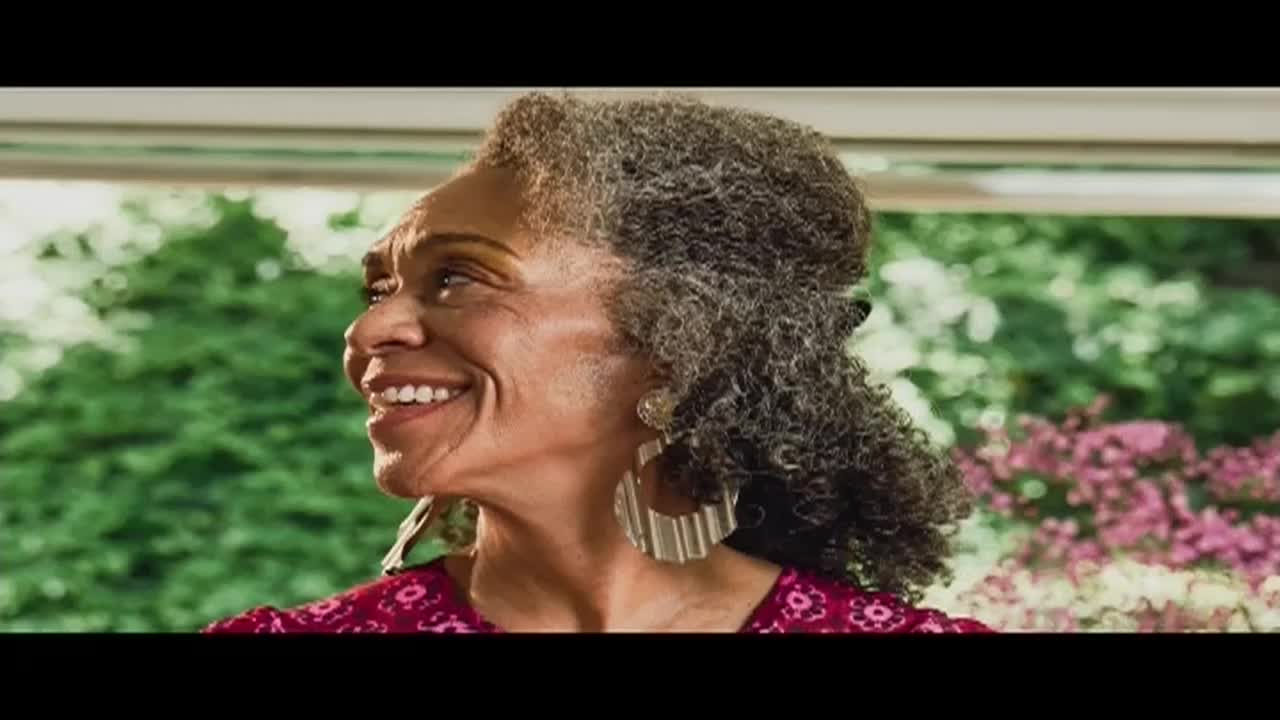 A woman with curly gray hair and large earrings smiles, looking off to the side. Her red patterned top is visible, and a blurred green and pink garden provides a backdrop.
