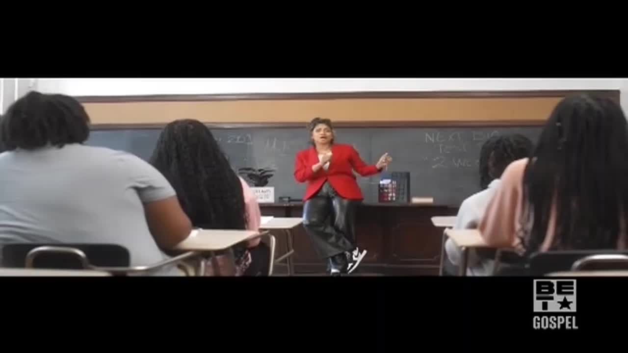 A woman in a red blazer stands at the front of a classroom, gesturing with her hands. Students sit at their desks, facing forward, as if listening to a lecture.
