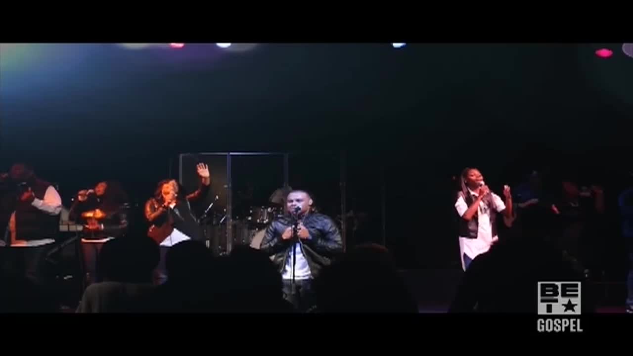 The singers on stage are animated, some with their hands raised, others clutching microphones. The BET Gospel logo is visible, and the crowd is dark and indistinct.

