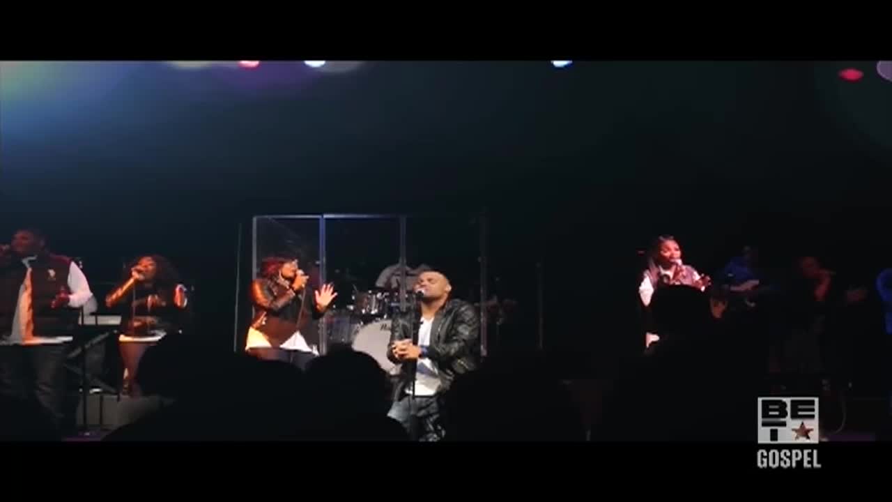 The lead singer, in a leather jacket, stands center stage, eyes closed, hands clasped.  Behind him, a choir of singers in dark outfits belt out a song under the stage lights.
